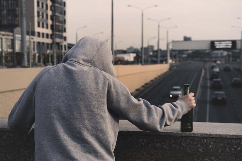 Mężczyzna w kapturze na głowie, twarz niewidoczna, trzyam w ręce butelkę alkoholu